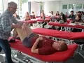 Un professeur pratique un examen de genou sur un étudiant allongé sur une table de massage rouge, dans une salle de classe d'une école de kinésithérapie suisse.