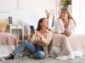 Deux étudiantes françaises discutent de leur aide au logement en Espagne, l'une sur le lit, l'autre assise au sol avec un téléphone.