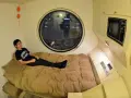 Un jeune homme se détend dans un lit, dans un petit logement étudiant à Tokyo. Une fenêtre ronde offre une vue nocturne sur la ville.