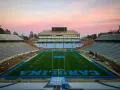 Stade de football vide de l'université de Caroline du Nord à Chapel Hill, avec des sièges bleus et le logo des Tar Heels. Le ciel rose du crépuscule surplombe le terrain.