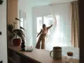Une jeune femme aux cheveux blonds dénoue les rideaux d'une chambre lumineuse. Un aperçu de la vie étudiante à Genève, où le logement étudiant et son prix sont une préoccupation majeure.