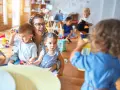 Une enseignante souriante, en formation **master petite enfance à distance**, interagit avec des enfants dans une salle de jeux colorée.