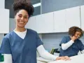 Une femme souriante en uniforme médical, prête pour sa formation secrétaire médicale en ligne. Une collègue au téléphone en arrière-plan.