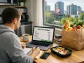 Un homme analyse son budget mensuel sur son ordinateur portable, comparant le coût de la vie au Canada. Une épicerie est à côté.