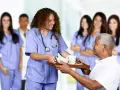 Une jeune femme en formation paramédicale sans bac, souriante, sert un plateau repas à un patient.