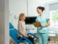 Une femme en blouse médicale parle à une patiente assise sur un fauteuil d'examen bleu.