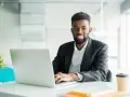 Un homme souriant travaille sur son ordinateur portable, peut-être pour son msc finance online. Il est dans un bureau moderne.