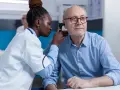 Une jeune femme en formation BTS Audioprothésiste examine l'oreille d'un patient âgé avec un otoscope.