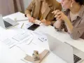 Deux femmes analysent des documents financiers et utilisent une calculatrice. Leurs mains pointent vers des graphiques, suggérant une discussion sur le pilotage de la performance, peut-être dans le cadre d'un msc audit à KEDGE.