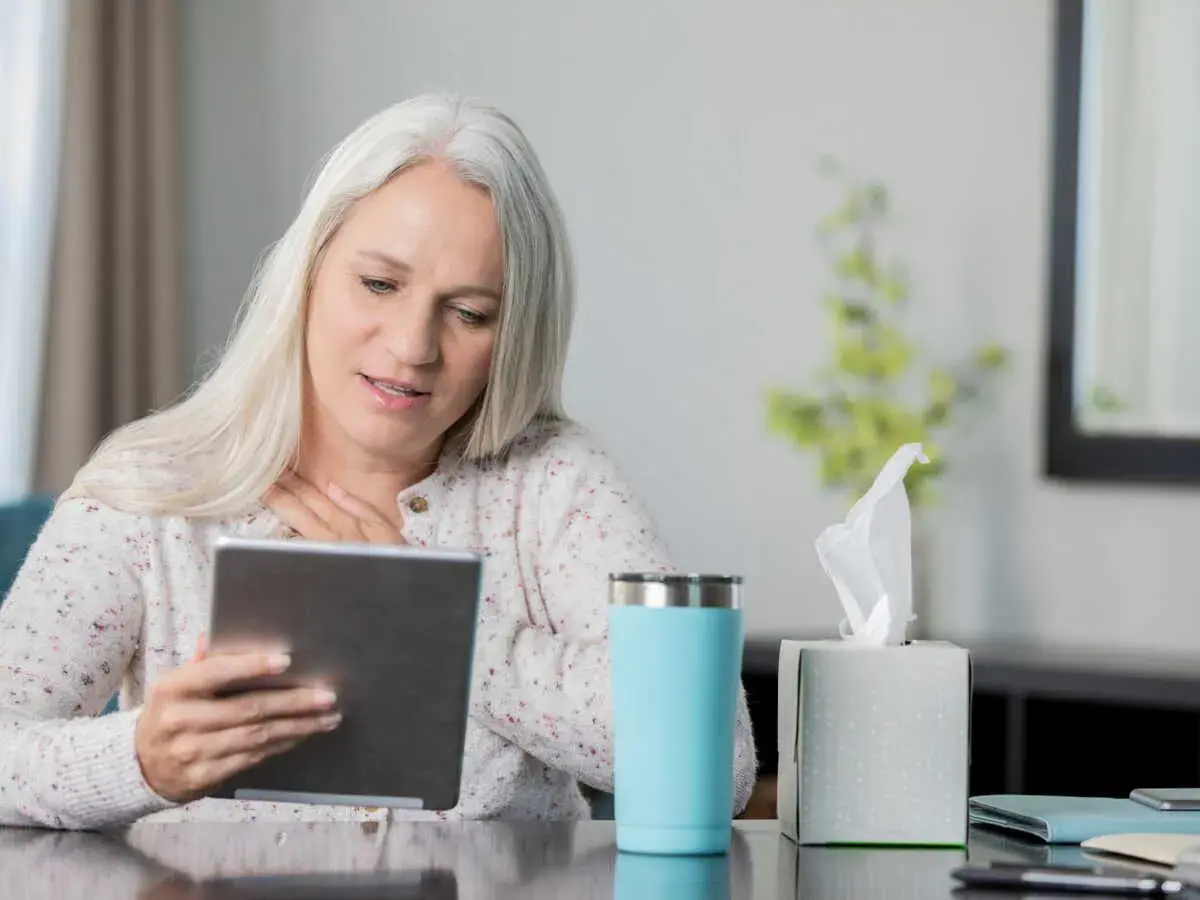 Femme aux cheveux gris suivant une formation orthophoniste à distance sur sa tablette, une tasse et des mouchoirs à proximité.