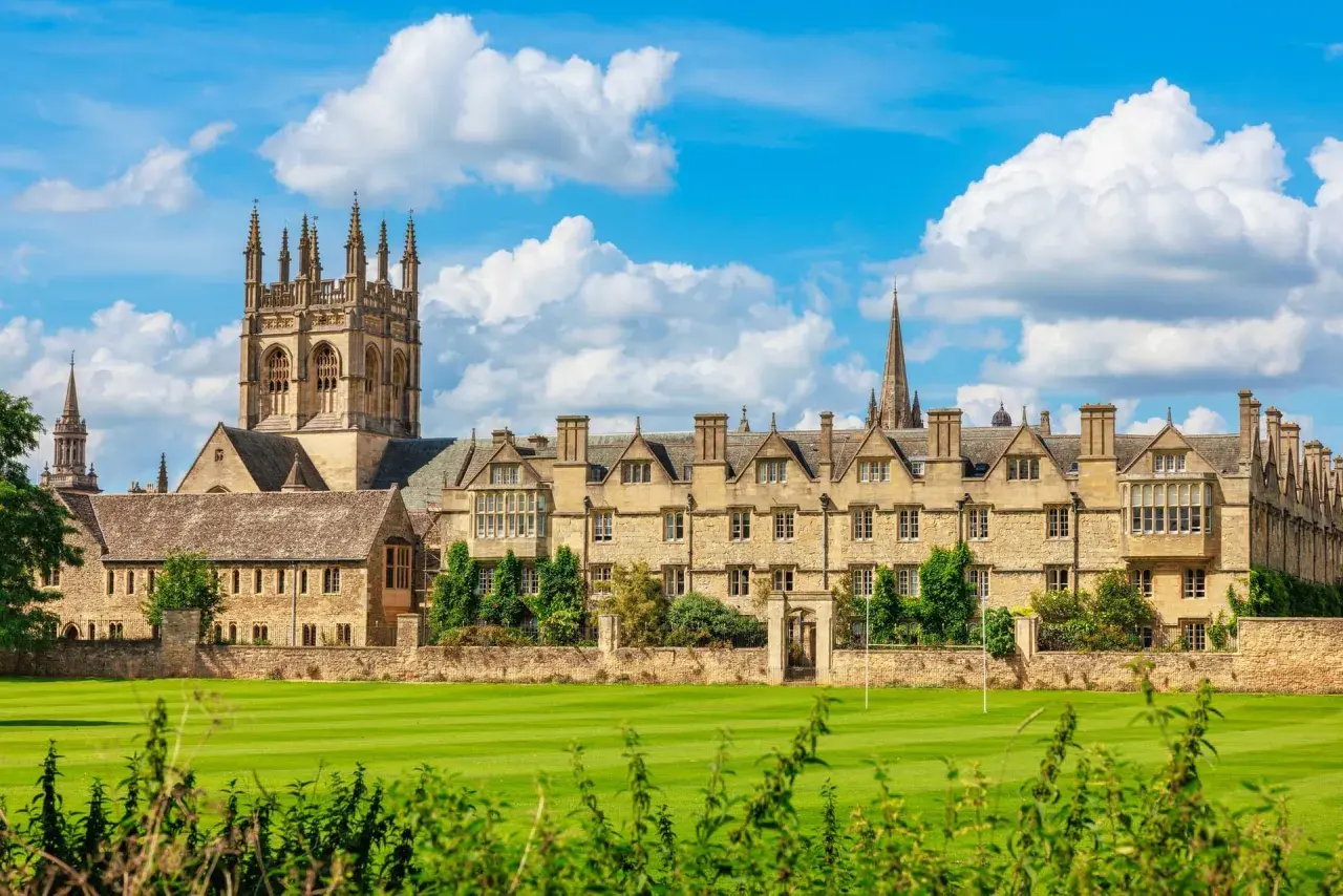 Magnifique vue sur l'université d'Oxford, avec ses bâtiments historiques en pierre, ses tours imposantes et son vaste terrain de jeu verdoyant sous un ciel bleu parsemé de nuages.