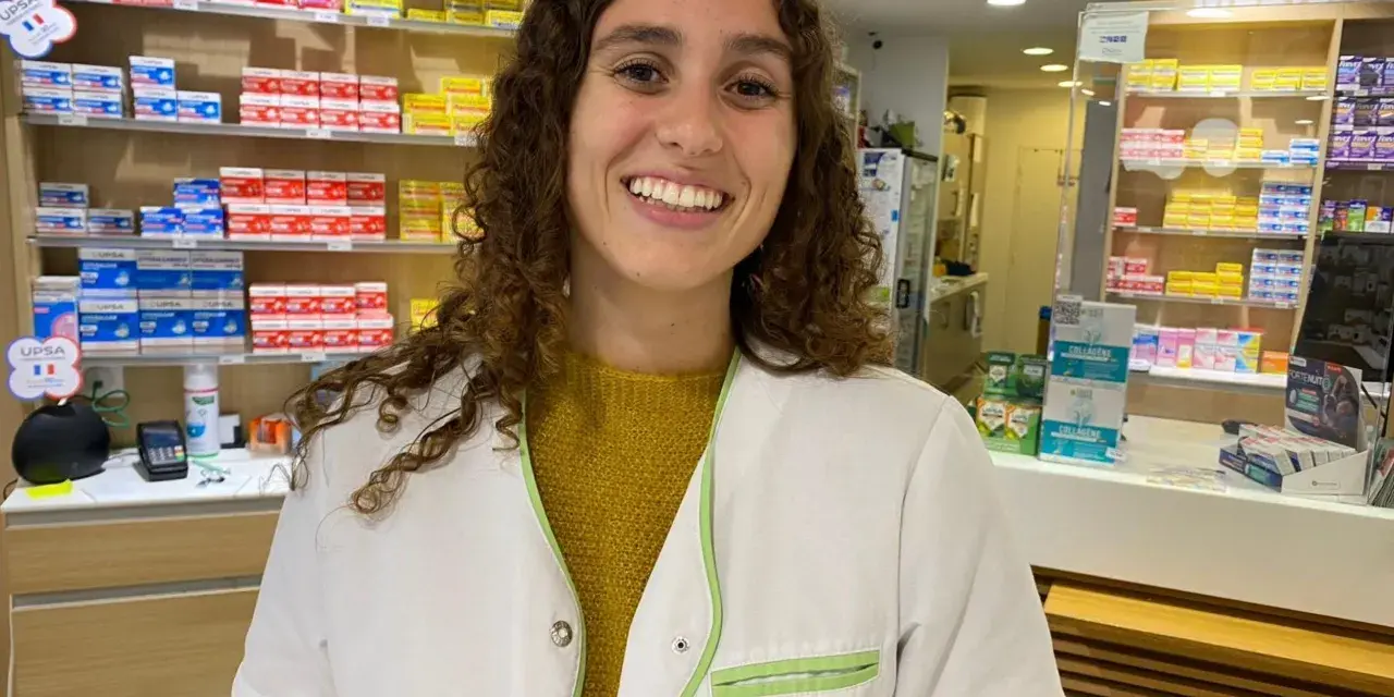 Une souriante préparatrice en pharmacie en Espagne, entourée de boîtes de médicaments sur les étagères.