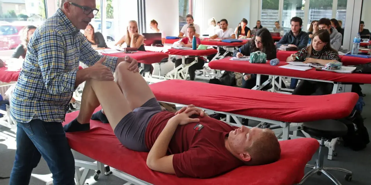 Un professeur pratique un examen de genou sur un étudiant allongé sur une table de massage rouge, dans une salle de classe d'une école de kinésithérapie suisse.