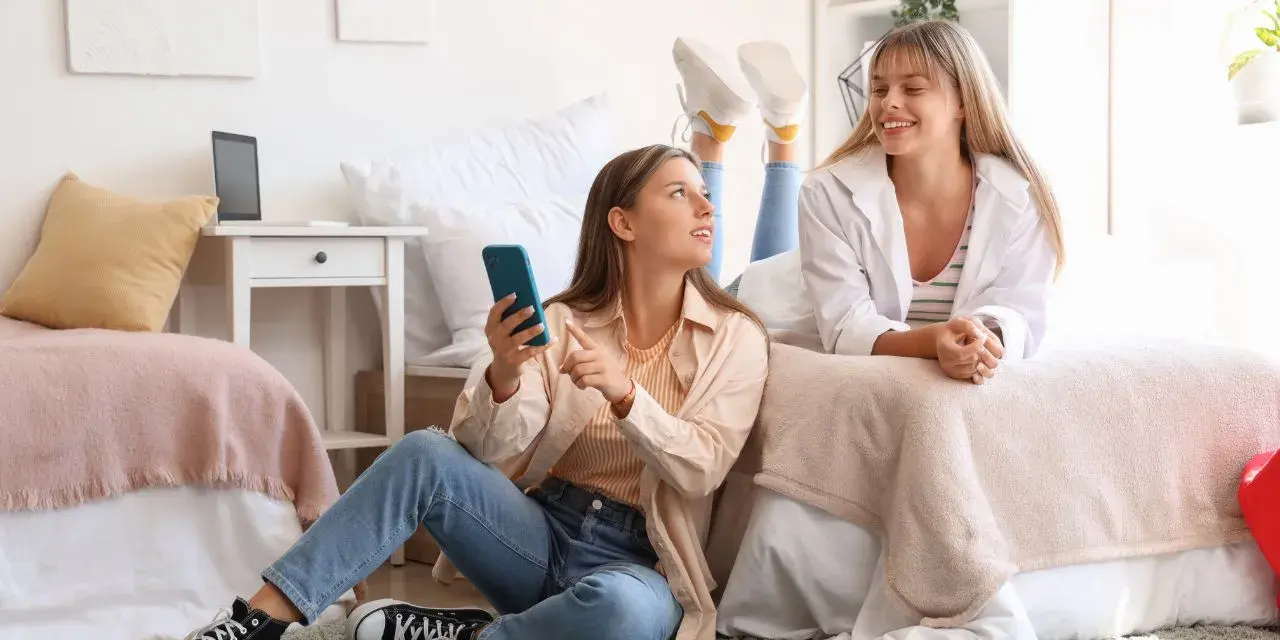 Deux étudiantes françaises discutent de leur aide au logement en Espagne, l'une sur le lit, l'autre assise au sol avec un téléphone.