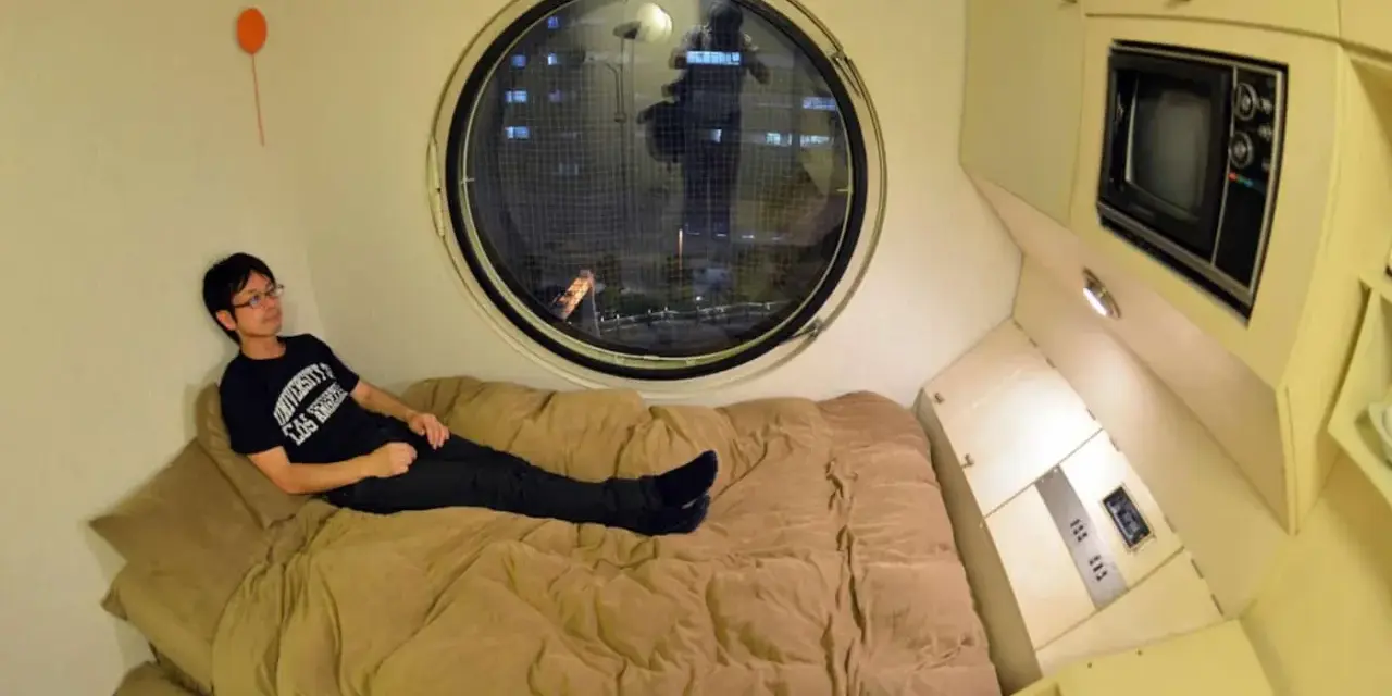Un jeune homme se détend dans un lit, dans un petit logement étudiant à Tokyo. Une fenêtre ronde offre une vue nocturne sur la ville.