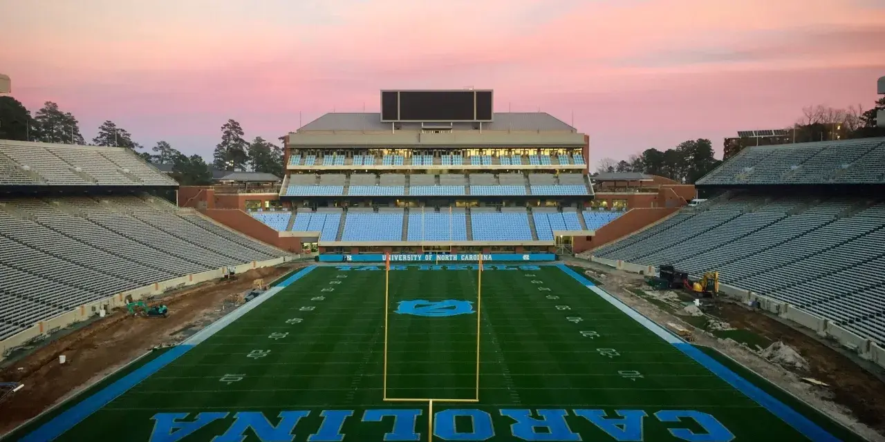 Stade de football vide de l'université de Caroline du Nord à Chapel Hill, avec des sièges bleus et le logo des Tar Heels. Le ciel rose du crépuscule surplombe le terrain.