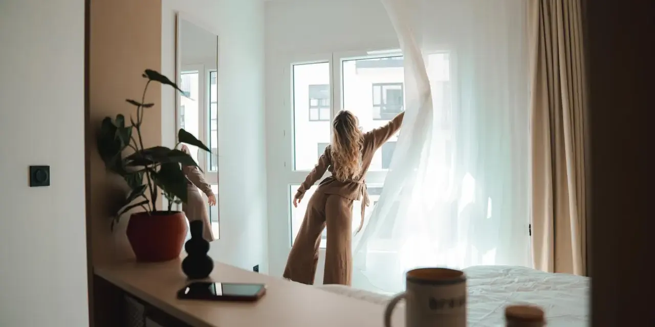 Une jeune femme aux cheveux blonds dénoue les rideaux d'une chambre lumineuse. Un aperçu de la vie étudiante à Genève, où le logement étudiant et son prix sont une préoccupation majeure.