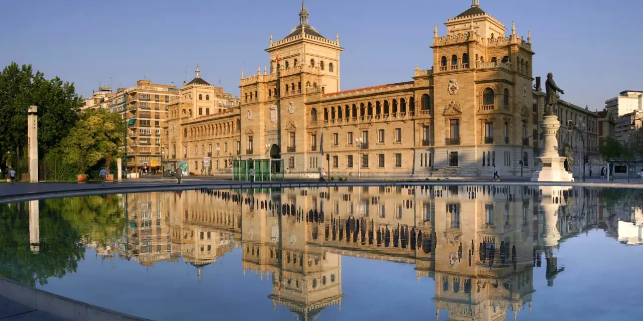 Le majestueux bâtiment du **universidad de valladolid** se reflète dans l'eau calme d'une fontaine, sous un ciel bleu clair.