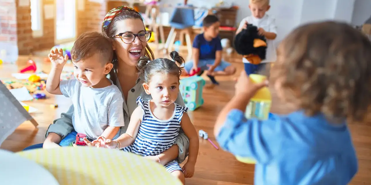 Une enseignante souriante, en formation **master petite enfance à distance**, interagit avec des enfants dans une salle de jeux colorée.