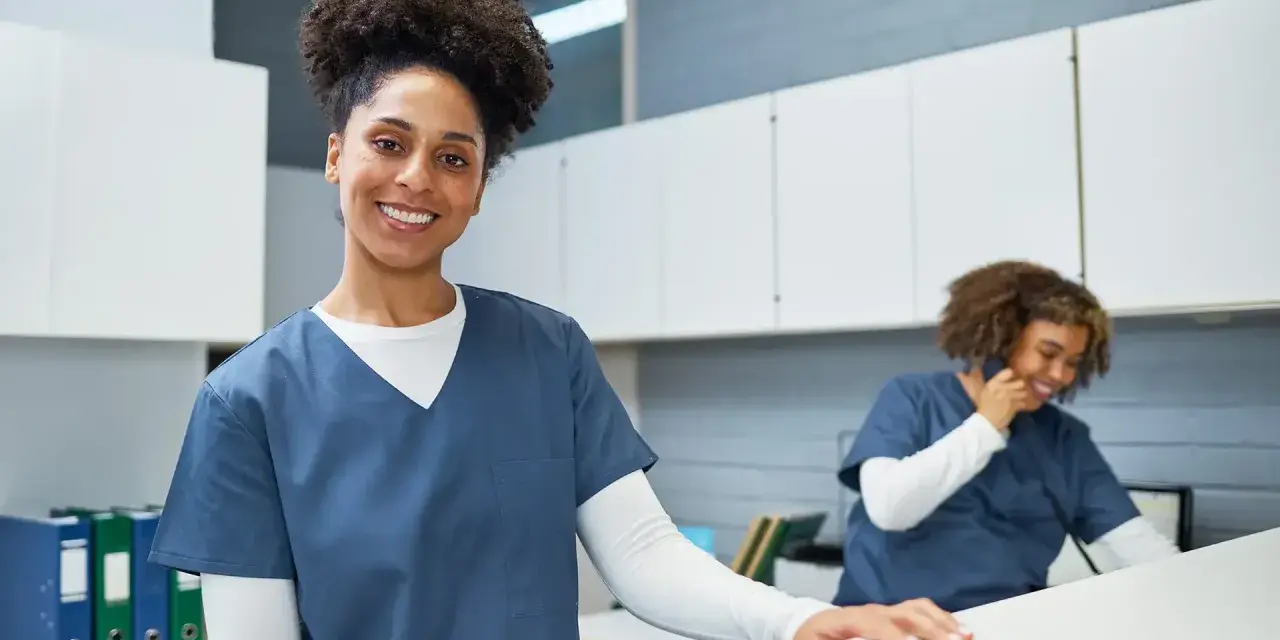 Une femme souriante en uniforme médical, prête pour sa formation secrétaire médicale en ligne. Une collègue au téléphone en arrière-plan.
