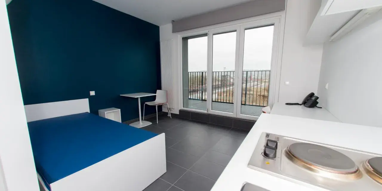 Chambre moderne pour étudiant à Luxembourg : lit bleu, bureau, kitchenette et balcon avec vue.