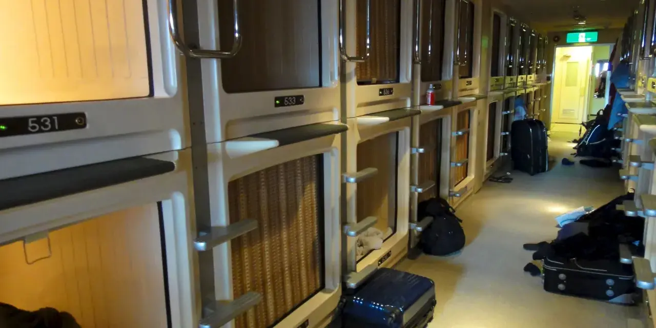 Couloir d'un hôtel capsule au Japon, avec des rangées de compartiments pour le **loyer au Japon**. Des valises sont posées au sol.