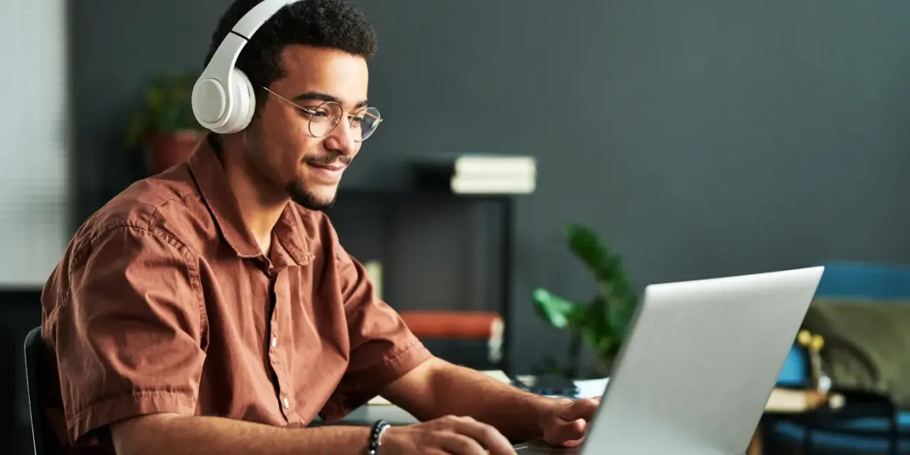 Jeune homme souriant avec des écouteurs, concentré sur son ordinateur portable. Il suit un master informatique à distance, profitant de la flexibilité de l'apprentissage en ligne.