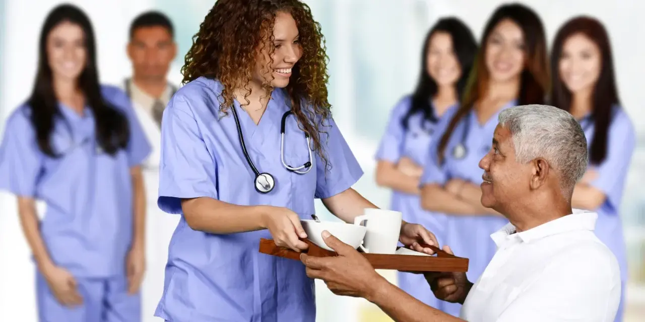 Une jeune femme en formation paramédicale sans bac, souriante, sert un plateau repas à un patient.
