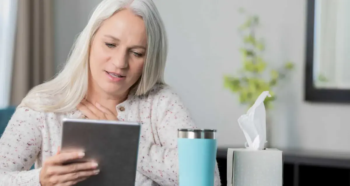 Femme aux cheveux gris suivant une formation orthophoniste à distance sur sa tablette, une tasse et des mouchoirs à proximité.