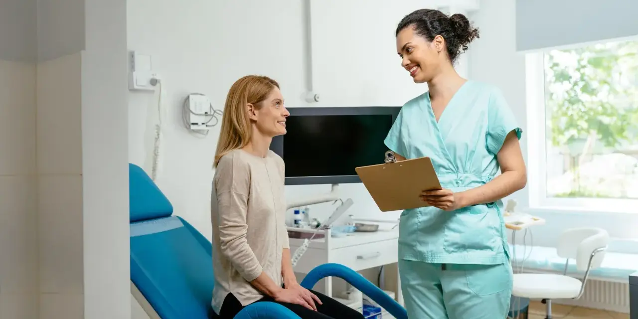 Une femme en blouse médicale parle à une patiente assise sur un fauteuil d'examen bleu.