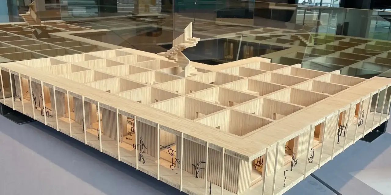 Maquette en bois d'une école d'architecture belge, avec des figures stylisées et des plantes.