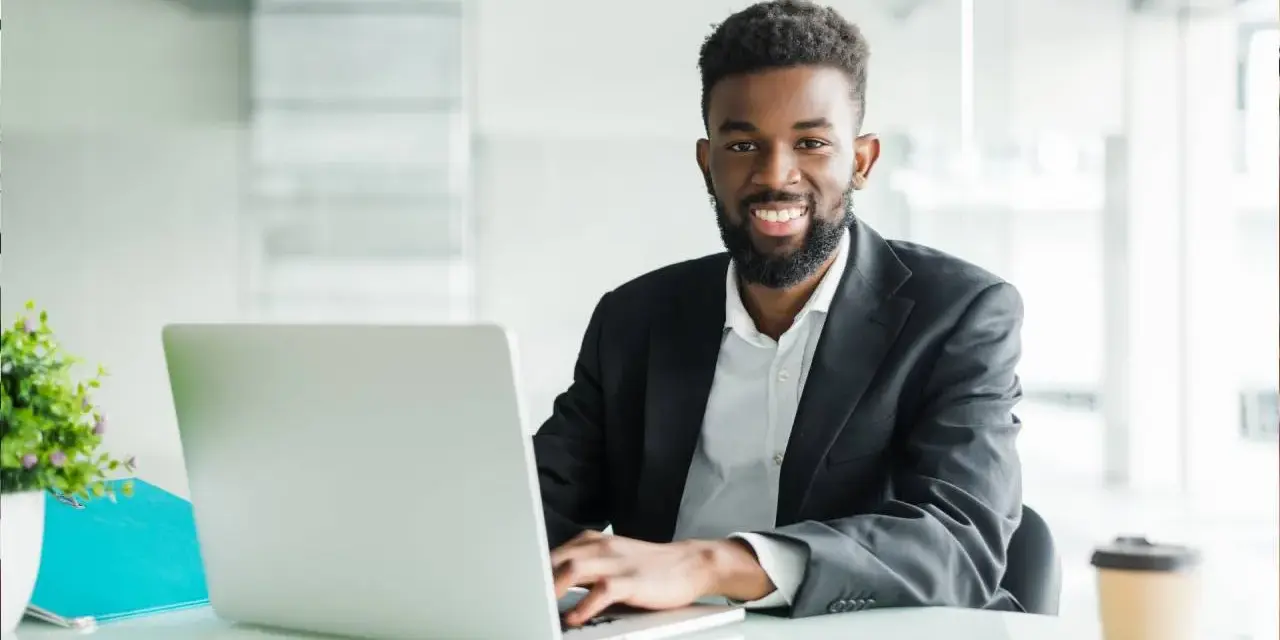 Un homme souriant travaille sur son ordinateur portable, peut-être pour son msc finance online. Il est dans un bureau moderne.