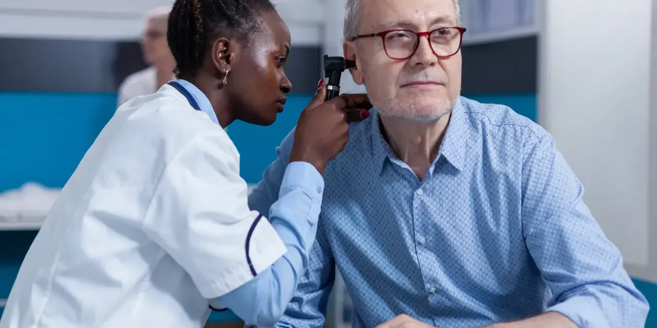 Une jeune femme en formation BTS Audioprothésiste examine l'oreille d'un patient âgé avec un otoscope.