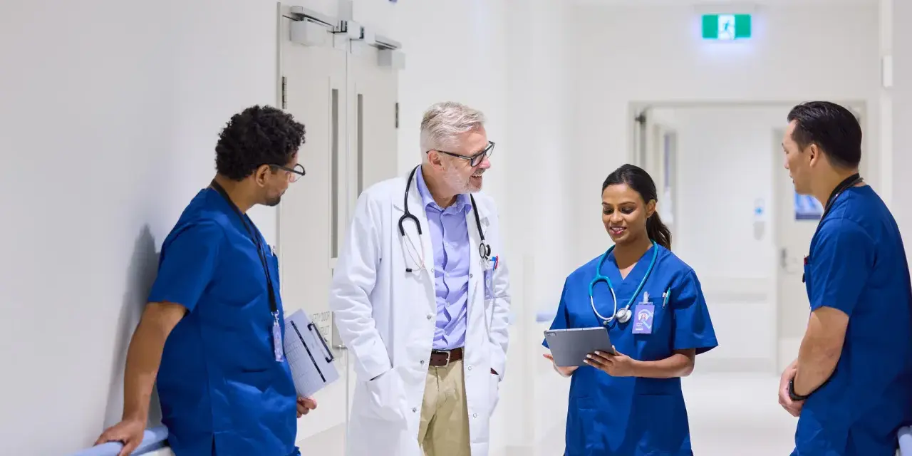 Une équipe médicale discute dans un couloir d'hôpital. Un médecin en blouse blanche échange avec trois infirmiers en uniforme bleu. Ils semblent discuter d'un cas, peut-être en lien avec du infirmier en ligne.
