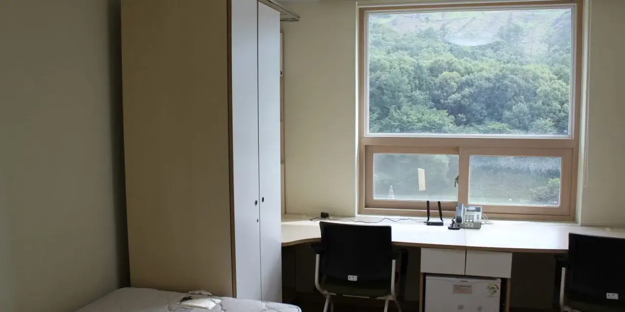 Chambre simple pour un logement étudiant à Séoul, avec un lit, une armoire, un bureau avec mini-frigo et une vue sur la verdure.