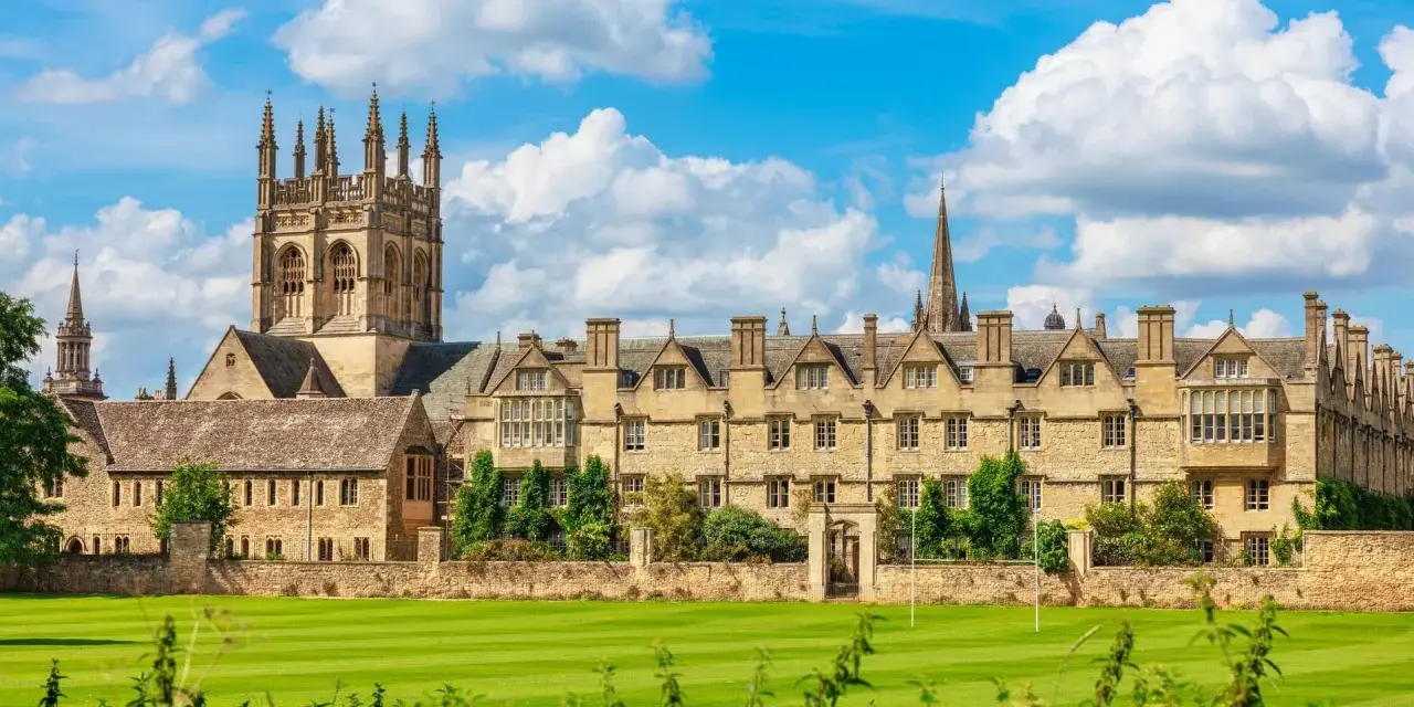 Magnifique vue sur l'université d'Oxford, avec ses bâtiments historiques en pierre, ses tours imposantes et son vaste terrain de jeu verdoyant sous un ciel bleu parsemé de nuages.
