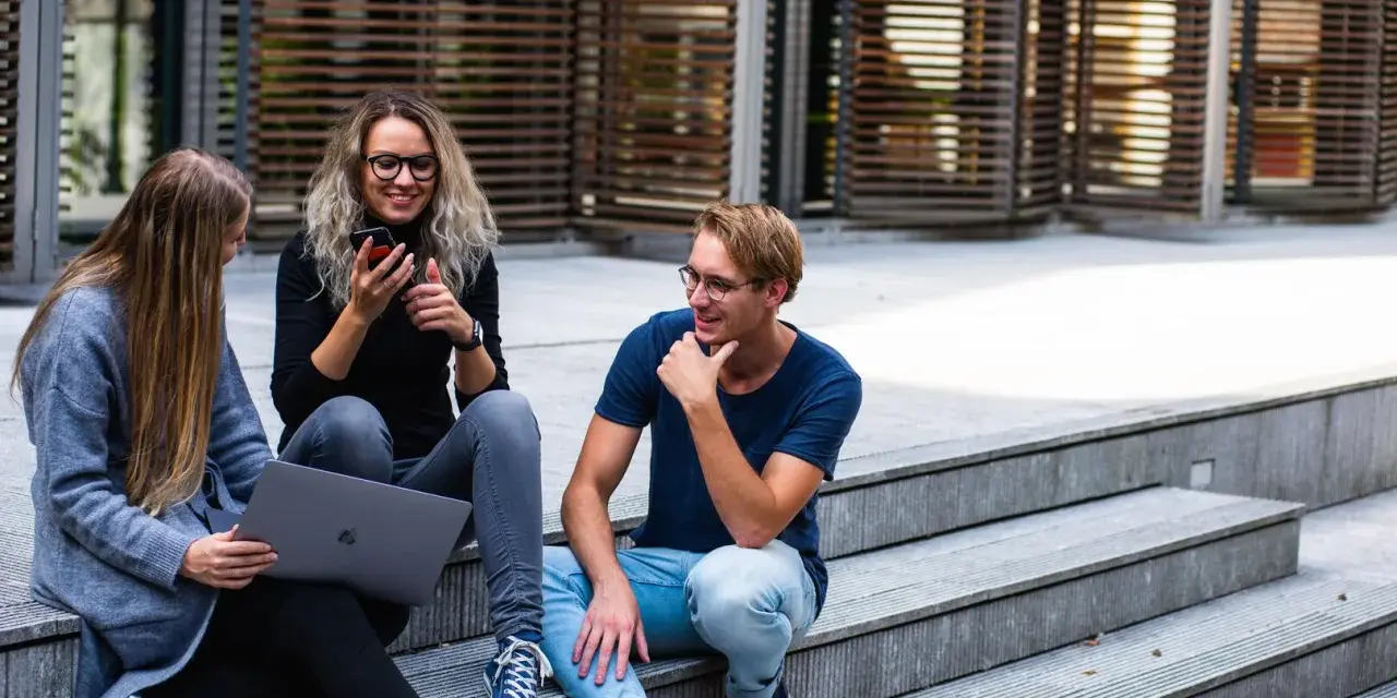 Trois jeunes discutent sur des marches, l'un regarde son téléphone, l'autre un ordinateur portable. Ils semblent parler du cout de la vie aux Pays-Bas.
