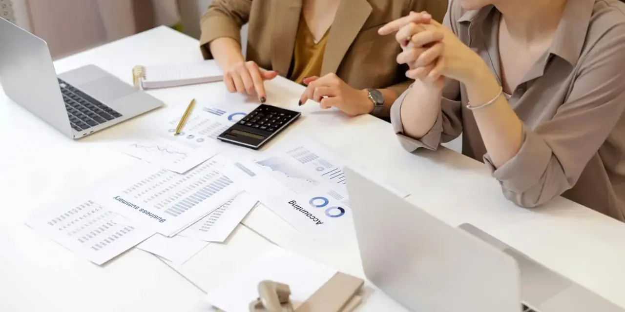 Deux femmes analysent des documents financiers et utilisent une calculatrice. Leurs mains pointent vers des graphiques, suggérant une discussion sur le pilotage de la performance, peut-être dans le cadre d'un msc audit à KEDGE.