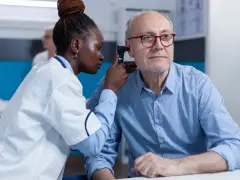 Une jeune femme en formation BTS Audioprothésiste examine l'oreille d'un patient âgé avec un otoscope.