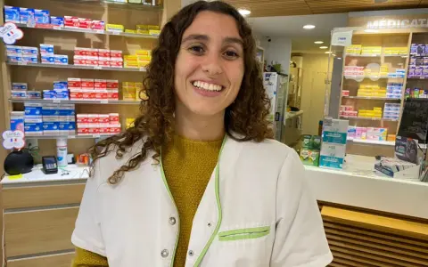 Une souriante préparatrice en pharmacie en Espagne, entourée de boîtes de médicaments sur les étagères.