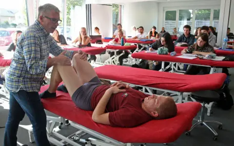 Un professeur pratique un examen de genou sur un étudiant allongé sur une table de massage rouge, dans une salle de classe d'une école de kinésithérapie suisse.