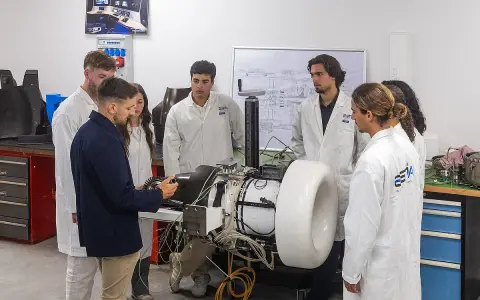 Des étudiants en blouse blanche observent un moteur complexe dans un laboratoire d'école d'ingénieur en Belgique.