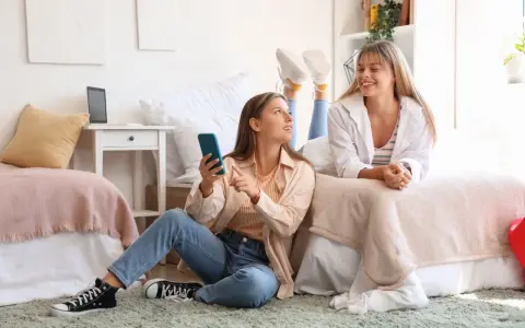 Deux étudiantes françaises discutent de leur aide au logement en Espagne, l'une sur le lit, l'autre assise au sol avec un téléphone.