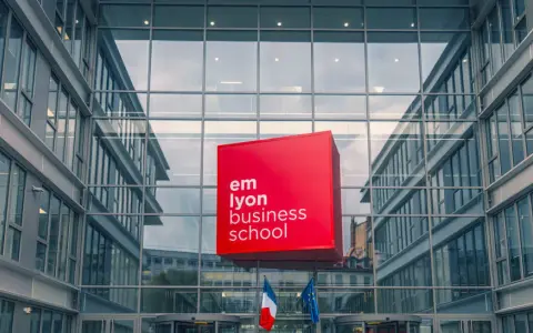 Le logo rouge d'EM Lyon Business School flotte devant un bâtiment moderne en verre. Idéal pour un master project management.