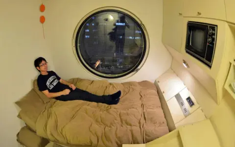 Un jeune homme se détend dans un lit, dans un petit logement étudiant à Tokyo. Une fenêtre ronde offre une vue nocturne sur la ville.