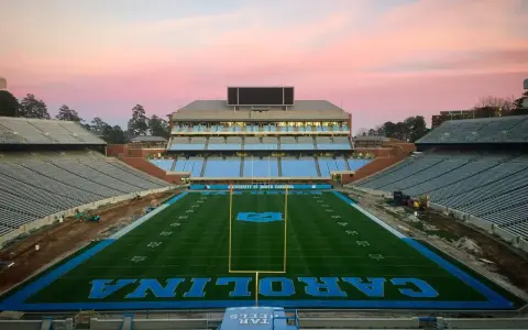 Stade de football vide de l'université de Caroline du Nord à Chapel Hill, avec des sièges bleus et le logo des Tar Heels. Le ciel rose du crépuscule surplombe le terrain.