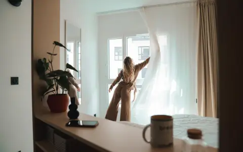 Une jeune femme aux cheveux blonds dénoue les rideaux d'une chambre lumineuse. Un aperçu de la vie étudiante à Genève, où le logement étudiant et son prix sont une préoccupation majeure.