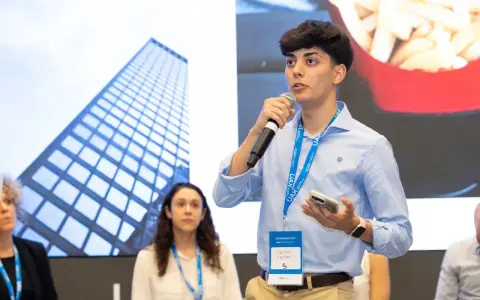 Un jeune homme prend la parole au micro, portant un badge UAX Madrid. Derrière lui, une image de gratte-ciel et des frites.