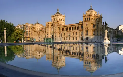 Le majestueux bâtiment du **universidad de valladolid** se reflète dans l'eau calme d'une fontaine, sous un ciel bleu clair.
