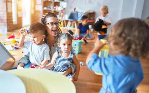 Une enseignante souriante, en formation **master petite enfance à distance**, interagit avec des enfants dans une salle de jeux colorée.